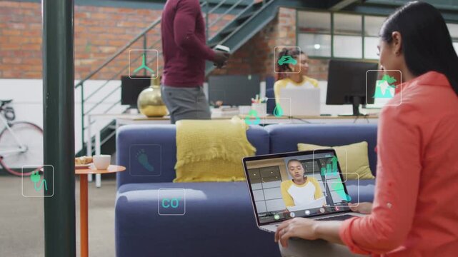 Woman operating laptop on remote call, peer walking by, green icons showing business data for team
