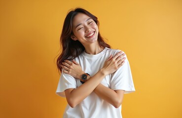 Obraz premium Young Asian woman smiles warmly, hugging herself in a studio with a bright yellow backdrop. She wears a simple white t-shirt and a watch, exuding positive self-love and inner peace.