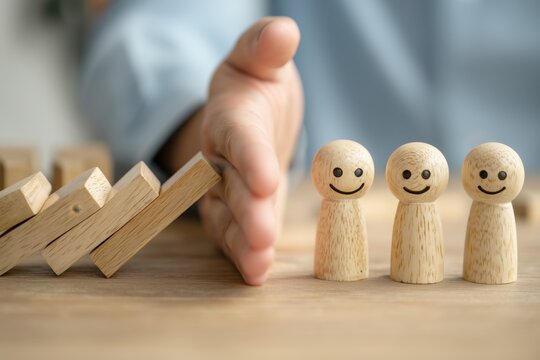 a close up view of a hand preventing a chain reaction of falling wooden dominoes from reaching three smiling wooden peg dolls on a light wooden surface Generative AI
