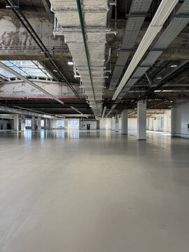 Spacious vacant warehouse interior featuring reflective epoxy flooring, ventilation ducts, pipes, and concrete pillars under industrial lighting.