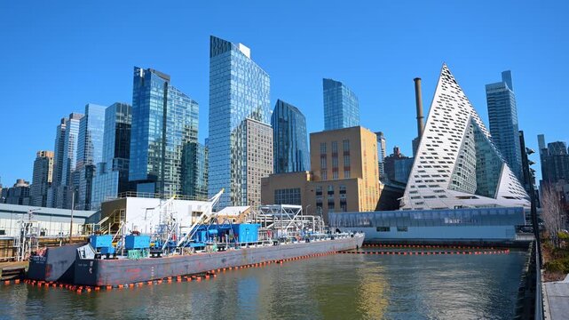 Industrial barge docked in Hudson River with Manhattan skyline background. Large industrial barge on water in front of modern architecture including VIA 57 West pyramid building in New York City.