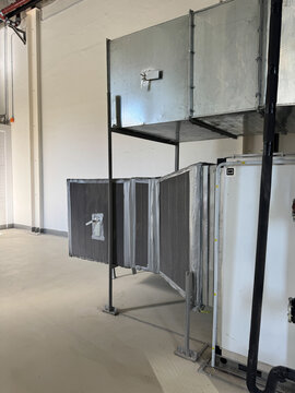 Large hvac air handling unit connected to rectangular metal ducts with grey thermal insulation. Ventilation system components installed in spacious industrial facility with concrete floor.