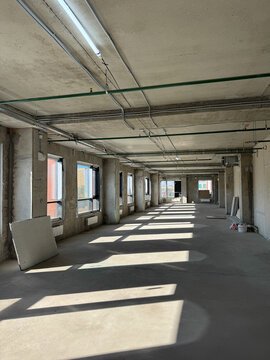 Spacious industrial room in shell and core state featuring raw concrete floor, large windows, green fire sprinkler pipes, and electrical wiring. Natural light creates shadows in unfinished interior.