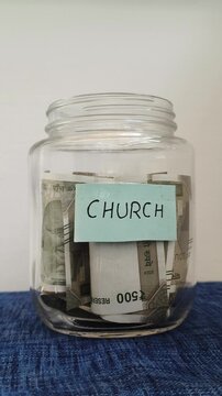 Hand putting india rupee currency note inside transparent glass jar container isolated on table with church word or text label. Saving money for church contribution or donation concept.