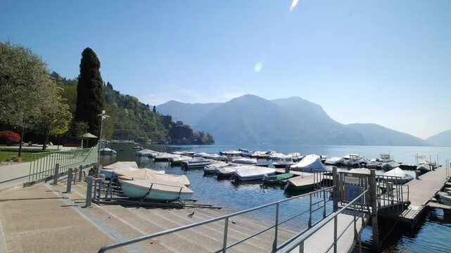 Wide shot of boats at Lake Lugano pier with scenic mountain peaks under a clear blue sky