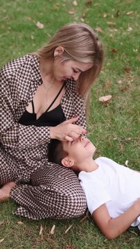 Vertical video. Loving parent. Family natural tenderness. Smiling young son resting on mother lap enjoying leisure together on green grass.