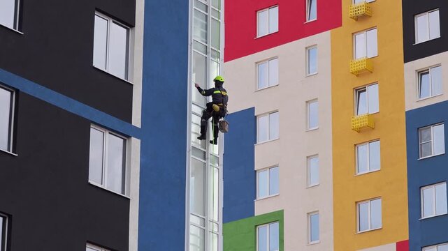 Industrial mountaineering man worker washing glazing using water squeegee, hanging from residential building. Male rope access laborer in work uniform during high-rise job. Industry urban works concep