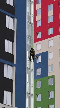Industrial mountaineering man worker washing glazing using water squeegee, hanging from residential building. Male rope access laborer in work uniform during high-rise job. Industry urban works concep