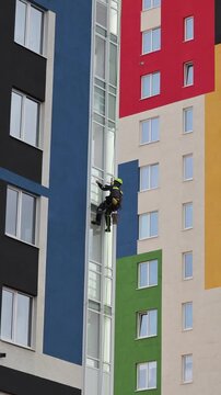 Industrial mountaineering man worker washing glazing using water squeegee, hanging from residential building. Male rope access laborer in work uniform during high-rise job. Industry urban works concep