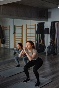 Women perform squats in a fitness studio with exercise mats and gym equipment in the background during a workout session