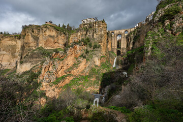 Fototapeta premium A traveler stands on a rock overlooking a waterfall and the massive arches of the Puente Nuevo bridge from the bottom of the gorge.