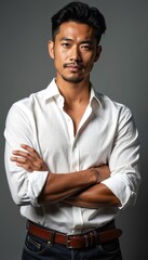 Obraz premium Man with crossed arms wears white shirt. He has neat hair and stubble. He looks seriously at camera. Dark jeans and brown belt complete his outfit. This portrait is suitable for fashion.
