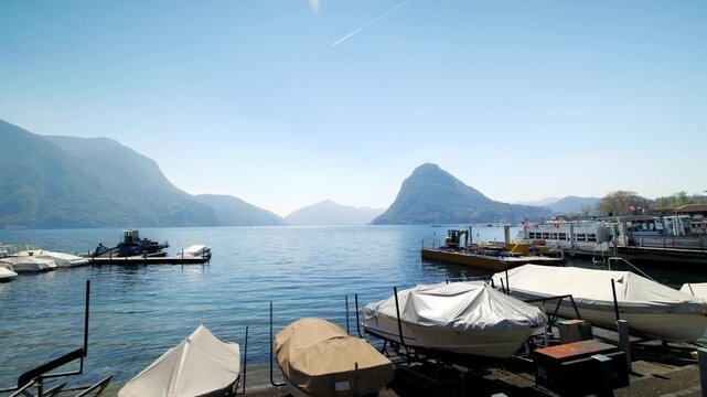 Panning shot of boats moored in Lugano marina with Lake Lugano and scenic mountains, Switzerland