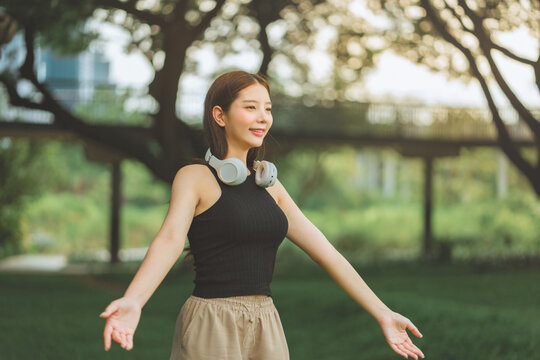 Happy Asian Gen Z woman with headphones standing in a green city park, spreading her arms wide to breathe fresh air. This joyful female adult enjoys an active lifestyle and mindfulness for wellness