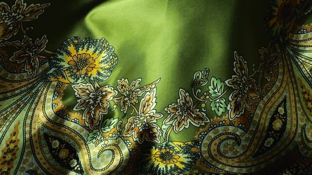 Green natural silk fabric with paisley and floral patterns waving in the wind. Flowing textile creates abstract waves and moving surface. Bright olive and golden colors of silky material.