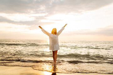 A woman stands by the shore with the ocean behind her. She has curly hair and laughs as the sun sets in the background, creating a warm glow around her. © maxbelchenko