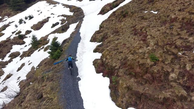 Drone follow shot from behind capturing a mountain biker climbing a steep road that narrows due to snow, eventually forcing the rider to stop and dismount