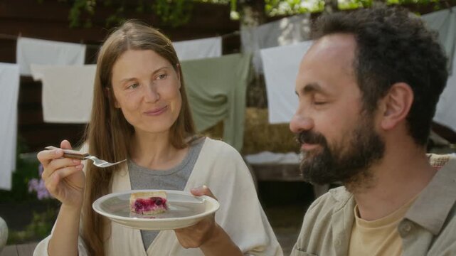 Medium close-up of smiling young Caucasian rural woman feeding husband piece of freshly baked organic cherry cheesecake, man enjoying and praising delicious dessert, during midday tea in backyard