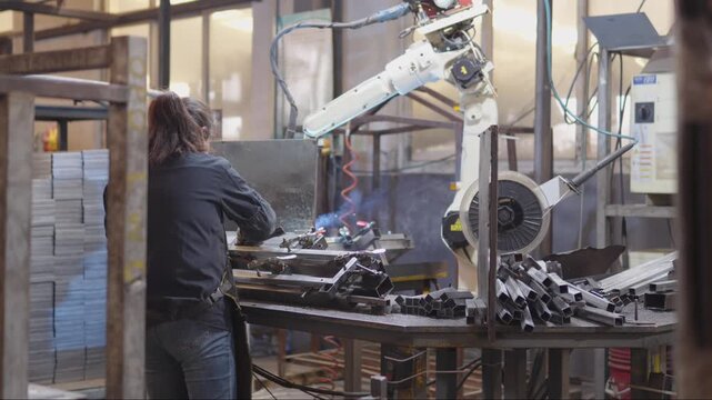 Industrial worker operating robotic arm to perform precision welding and grinding on metal components in a modern automated manufacturing facility, blending skill and technology