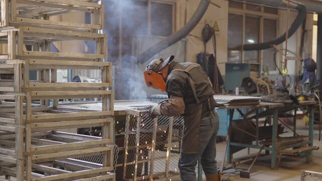 Industrial worker wearing protective gear using a welding torch to join metal pieces, generating bright sparks and smoke in busy manufacturing workshop