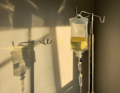 A glass intravenous drip bag holds yellow fluid. Medical equipment hangs on a stand, casting a shadow. Sunlight streams on the wall, suggesting a hospital room or clinic.