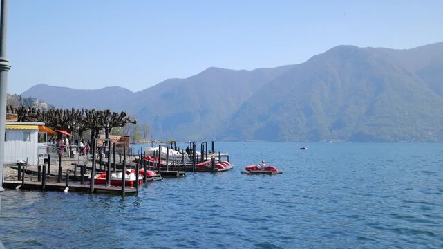 Static shot of Lake Lugano waterfront with red pedal boats at the pier and mountains, Switzerland