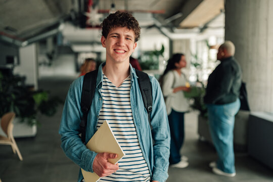 Young male student smiling at camera, holding book on modern university campus, embodying education and future success