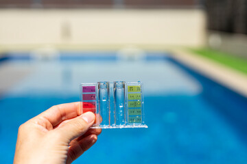 Close-up of a hand holding a pool water testing kit with color scale, measuring pH and chlorine levels in a clear blue swimming pool outdoors - pool maintenance concept © juancajuarez