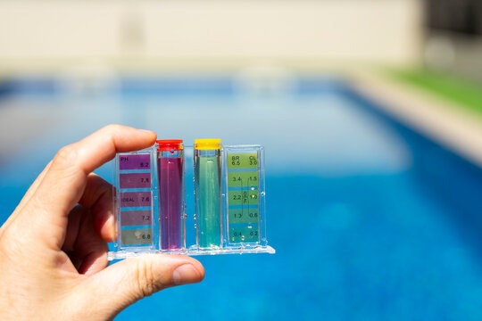 Close-up of a hand holding a pool water testing kit with color scale, measuring pH and chlorine levels in a clear blue swimming pool outdoors - pool maintenance concept