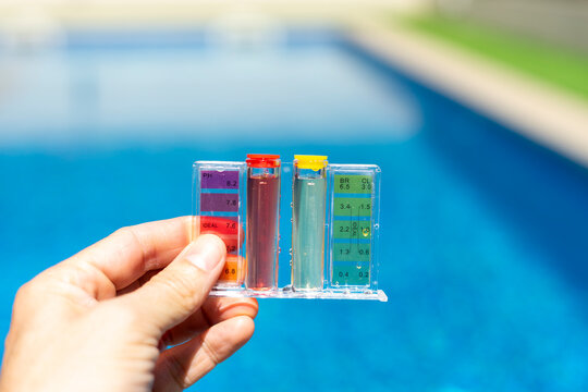 Close-up of a hand holding a pool water testing kit with color scale, measuring pH and chlorine levels in a clear blue swimming pool outdoors - pool maintenance concept