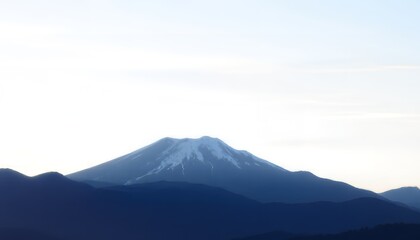 Fototapeta premium A snow-capped mountain stands tall against a clear blue sky with a range of dark mountains in the foreground at dusk or dawn.