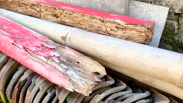 Old weathered wood planks with termite damage and peeling red paint stacked with bamboo and traditional clay roof tiles. Textures of decay, rustic materials, and architectural ruins.