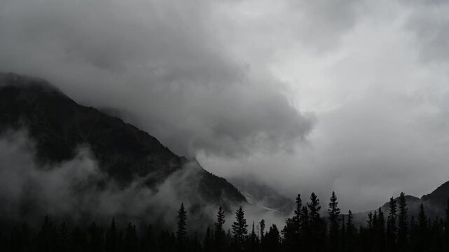 Timelapse of dramatic gray clouds swirling above silhouetted trees and rugged mountains in a dark moody wilderness landscape.