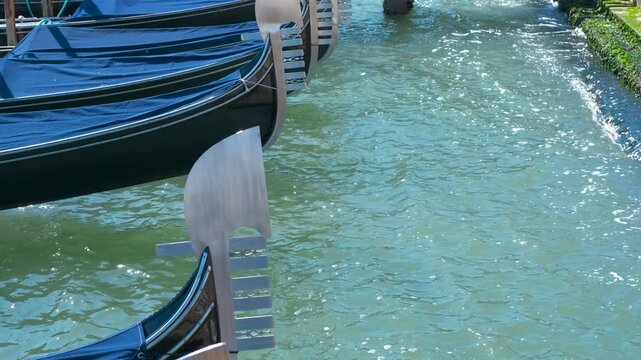 Gondolas moored in venice canal showing gentle water movement. Several gondolas, traditional venetian boats, are moored along a canal, their metallic prows gleaming in the sunligh