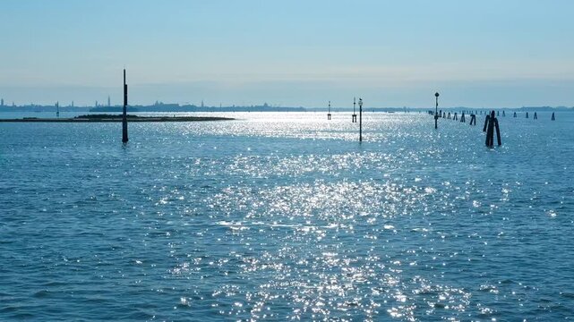 Sunlight reflecting on the venetian lagoon near venice. Bright sunlight reflects on the calm water surface of the venetian lagoon, with navigation poles and the silhouette of venice on the horizon