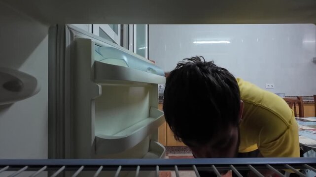 Frustrated man with a beard looking inside a completely empty refrigerator at night. Sad facial expression showing hunger and disappointment