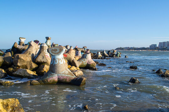 Concrete jacks painted with vibrant colors are positioned along a rocky shore, extending into the sea under a clear blue sky. The scene highlights the intertwining of art and nature.
