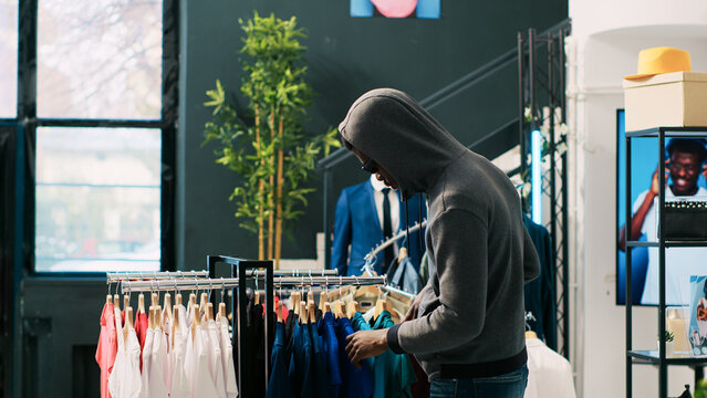 African american thief robbing clothing store, analyzing racks with stylish clothes. Robber trying to steal fashionable merchandise, wearing sunglasses and hood not to be recognized