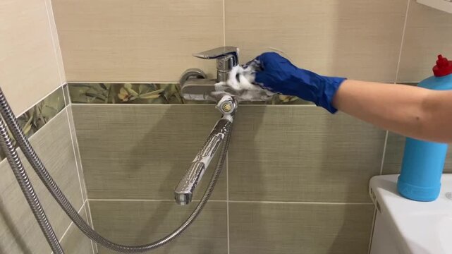 Blue gloved woman scrubbing chrome faucet with foam and sponge closeup of hand cleaning shower mixer, soap suds on tiles, flexible hose visible, careful rinse and polish for limescale removal