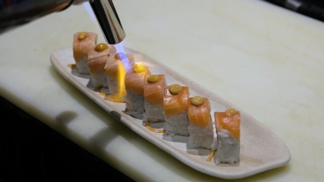 Dramatic close up of a professional chef using a culinary blowtorch to sear fresh salmon on top of uramaki sushi rolls creating a caramelized glaze and smoky flavor for a modern fusion Japanese dining