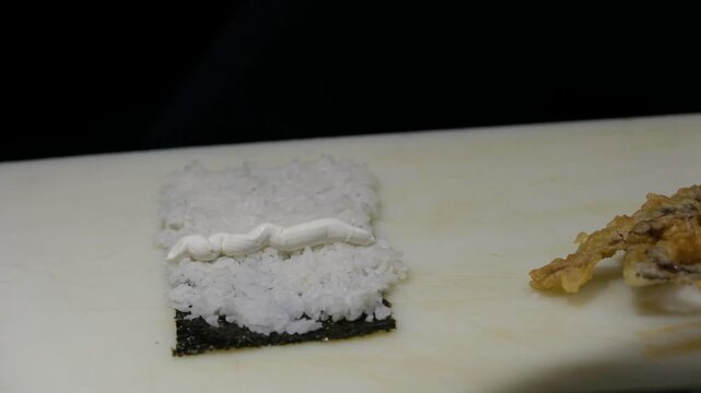 Professional sushi chef applying a thick layer of cream cheese over fresh white rice and nori seaweed sheet while preparing a gourmet Philadelphia maki roll in a clean Japanese restaurant kitchen