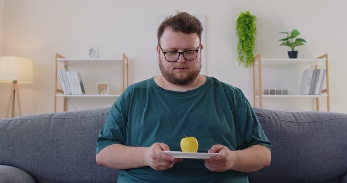Fat overweight man with glasses sits on a sofa studying an apple placed on a plate in a modern livingroom. Clear healthy choice for daily life. Focus on nutrition, diet, choice and lasting health.
