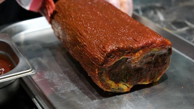 Extreme close up of a brush applying a red marinade over the textured surface of a roast meat roll in a professional kitchen emphasizing gourmet flavor and artisan cooking.