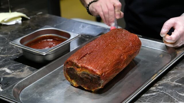 Close up of hands glazing a marinated beef roast with a red savory sauce in a professional kitchen environment representing gourmet food production and high-end catering service.