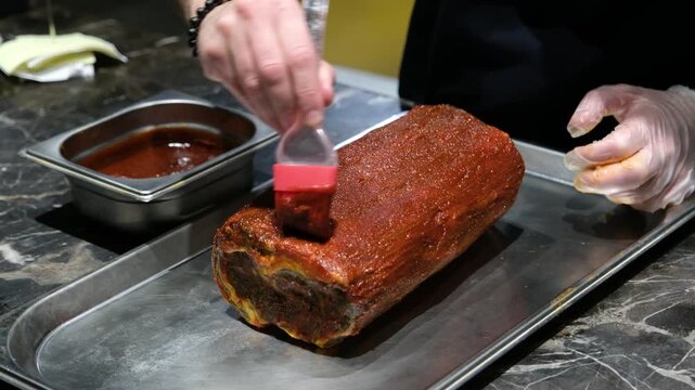 Professional chef using a brush to apply a rich glaze or sauce to a large meat roll on a metal tray in a commercial kitchen focusing on culinary techniques and food preparation.