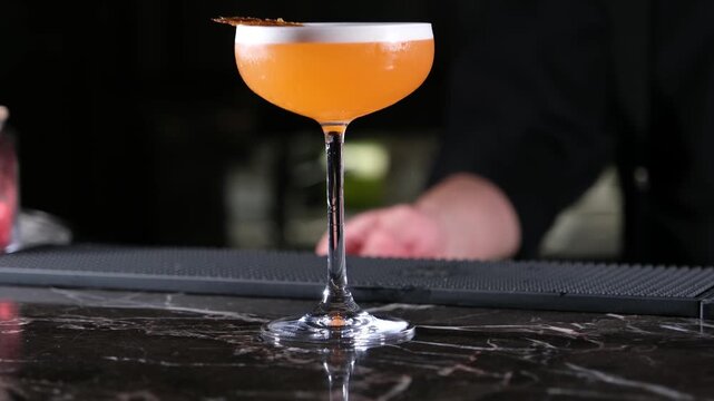 Professional cinematic shot of a colorful orange cocktail in a stem glass with artistic decoration on a dark marble counter representing gourmet beverage and fine dining lifestyle.