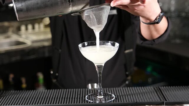 Professional bartender pouring a foamy white cocktail from a shaker through a double strainer into an elegant glass to create a smooth and frothy texture for a signature gourmet drink.