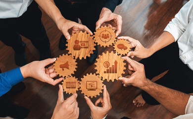 Diverse individuals join hands around wooden gears, representing teamwork and collaboration in a...