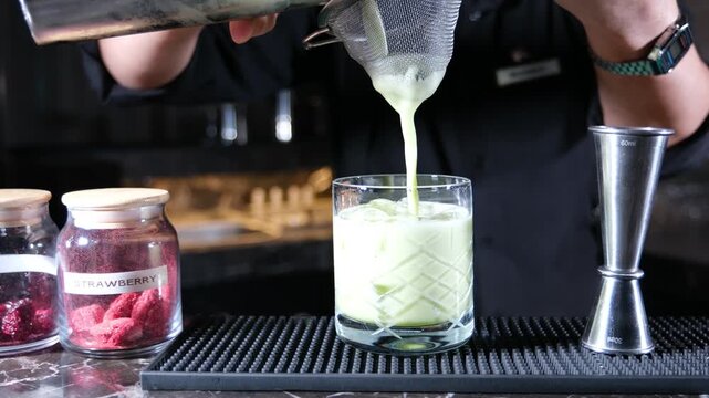 Professional bartender in a black uniform pouring a vibrant green tropical cocktail into a patterned rocks glass through a metal strainer during an evening hospitality service in a luxury lounge bar e