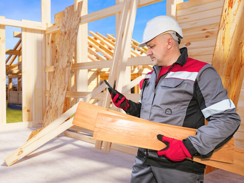 Timber construction, Wood frame assembly. Builder checks smartphone while carrying boards at house construction site, coordinating carpentry and material use.